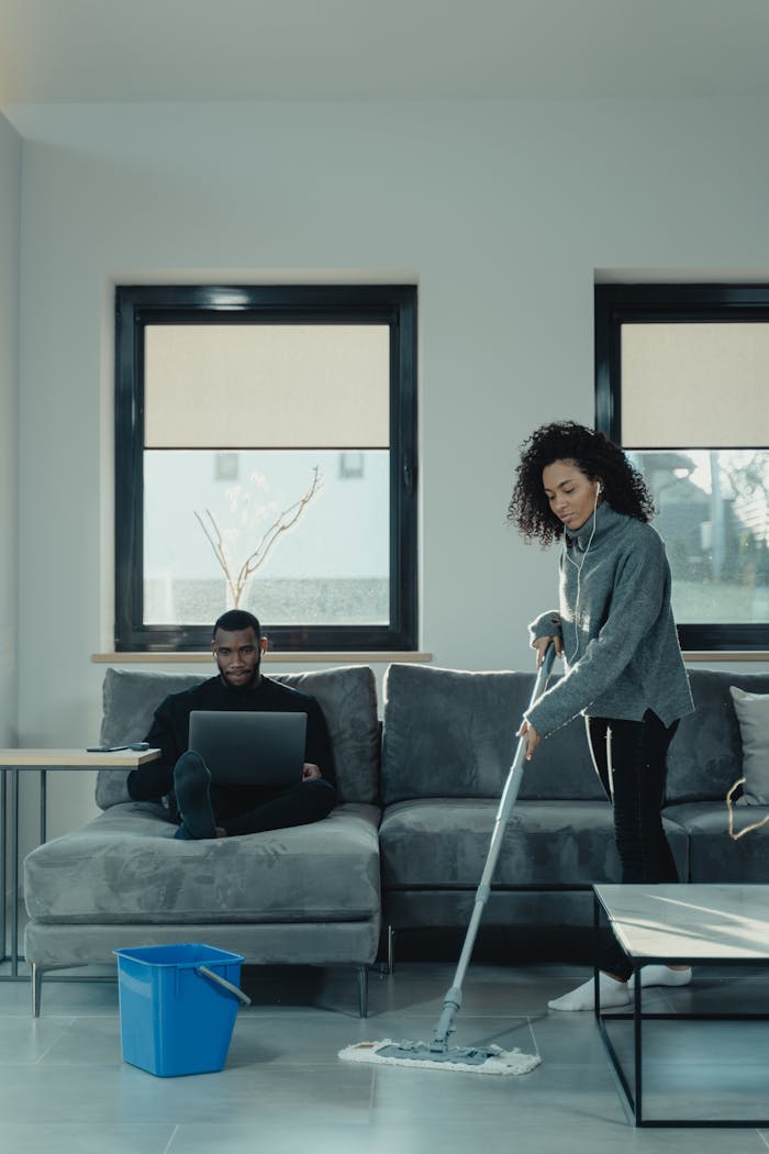 A man works on a laptop while a woman mops the floor in a cozy home setting.